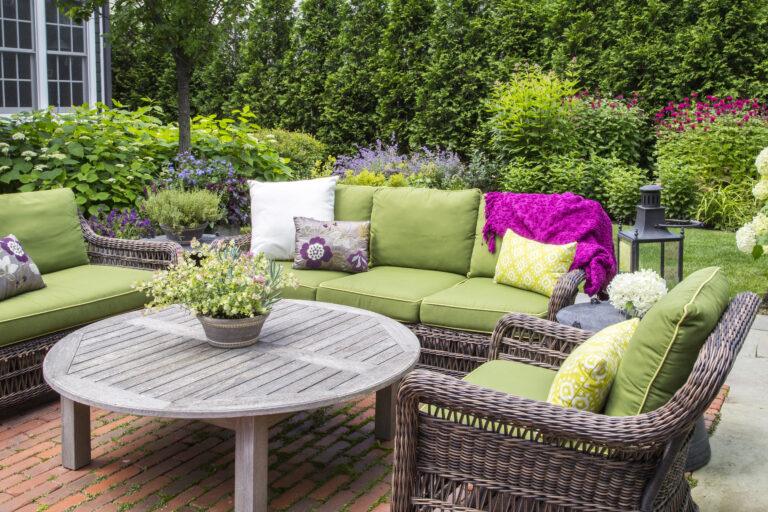 Wicker patio furniture with green cushions and patterned pillows surrounds a round wooden table, creating a cozy spot to enjoy lush plants and flowers—perfect inspiration for landscaping in Hinsdale, IL.