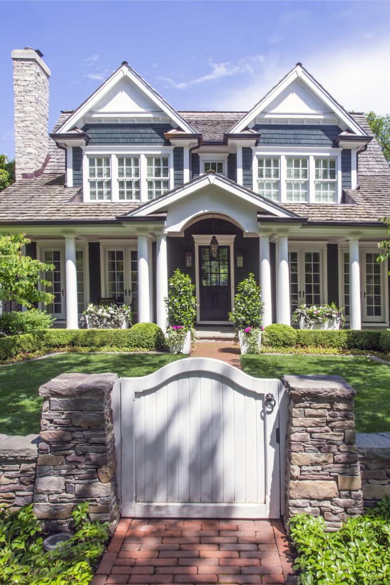 Two-story house with white columns, dark blue siding, and peaked roof, seen behind a stone wall and white gate, features expert landscaping in Hinsdale, IL for a beautifully manicured front yard.