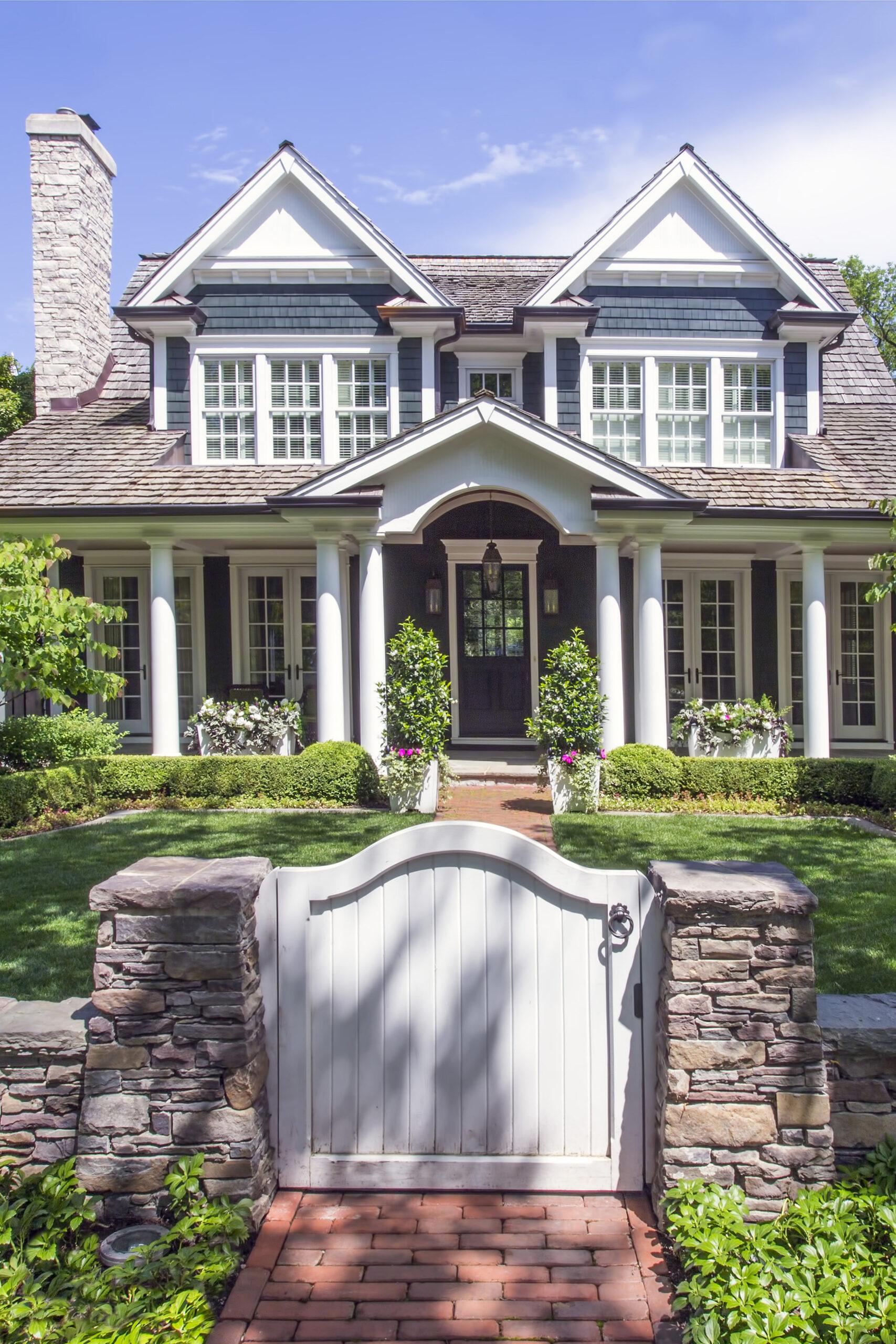 Two-story house with white columns, dark blue siding, and peaked roof, seen behind a stone wall and white gate, features expert landscaping in Hinsdale, IL for a beautifully manicured front yard.