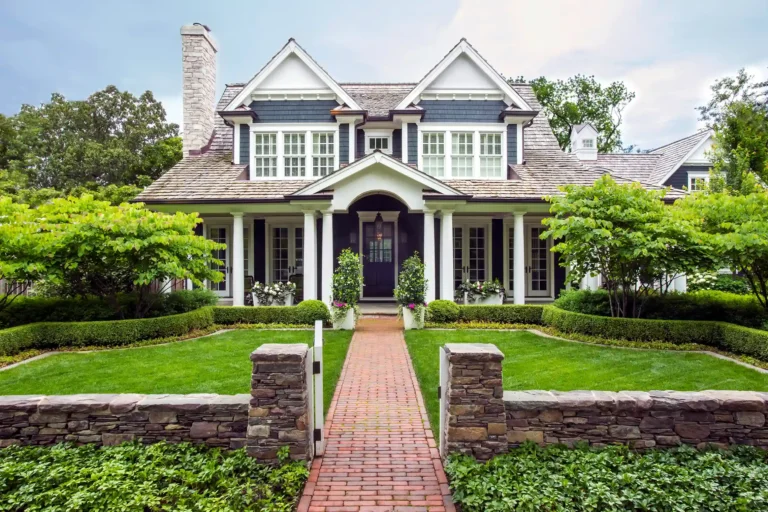A two-story suburban house with a front porch, white trim, manicured lawn, brick walkway, stone fence, and green trees on both sides.