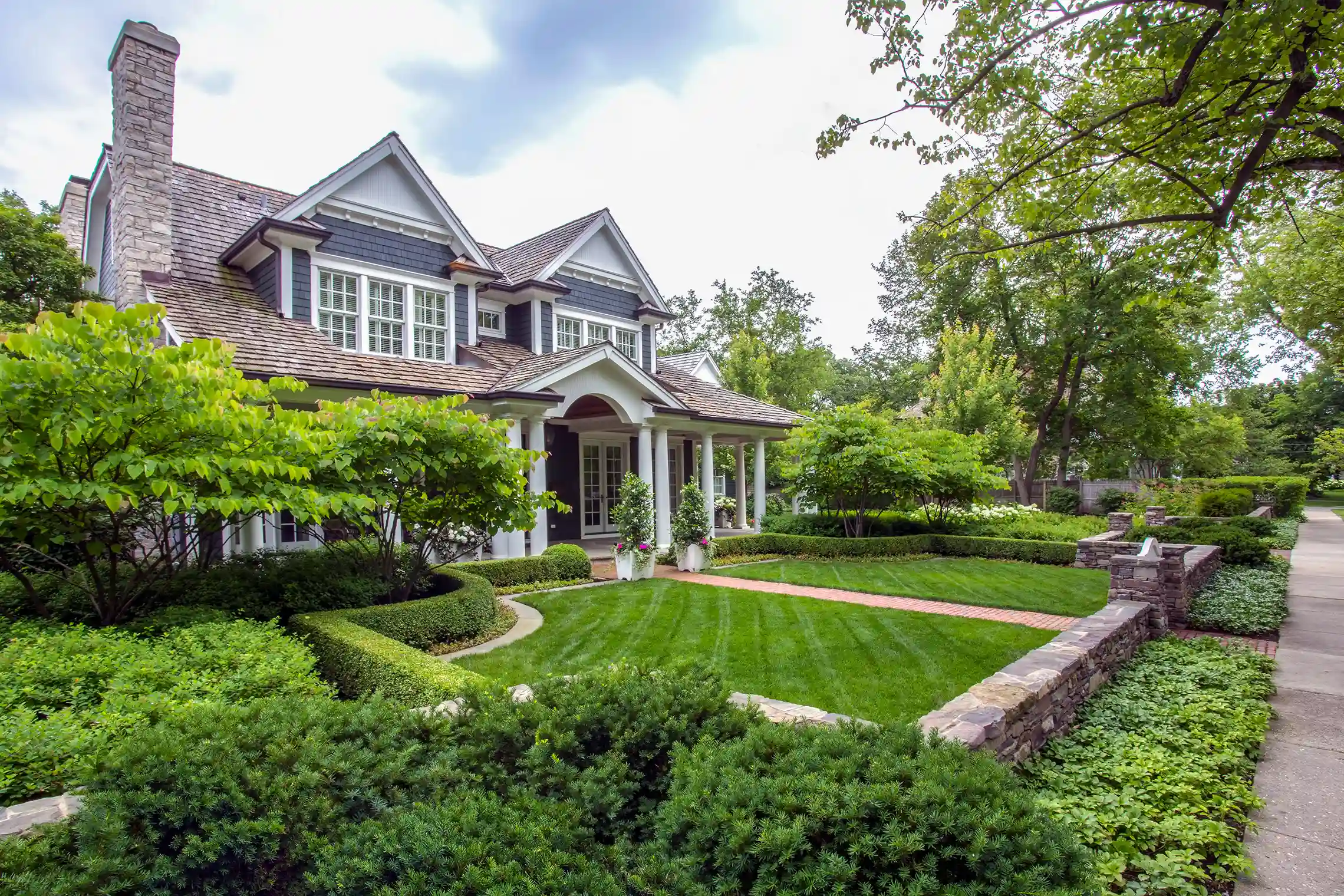 A two-story house with dark siding and white trim is surrounded by manicured hedges, green lawn, and mature trees along a stone-lined sidewalk.