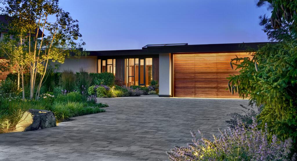 Modern single-story house with large wooden garage door, wide windows, and landscaped front yard at dusk.