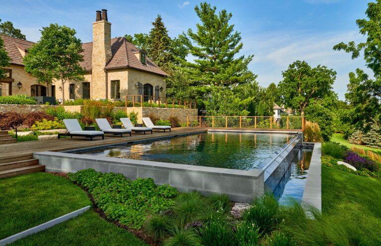 A modern infinity pool with several lounge chairs is surrounded by lush greenery, showcasing exquisite landscaping in Harbert, MI, and a stone house in the background under a clear blue sky.