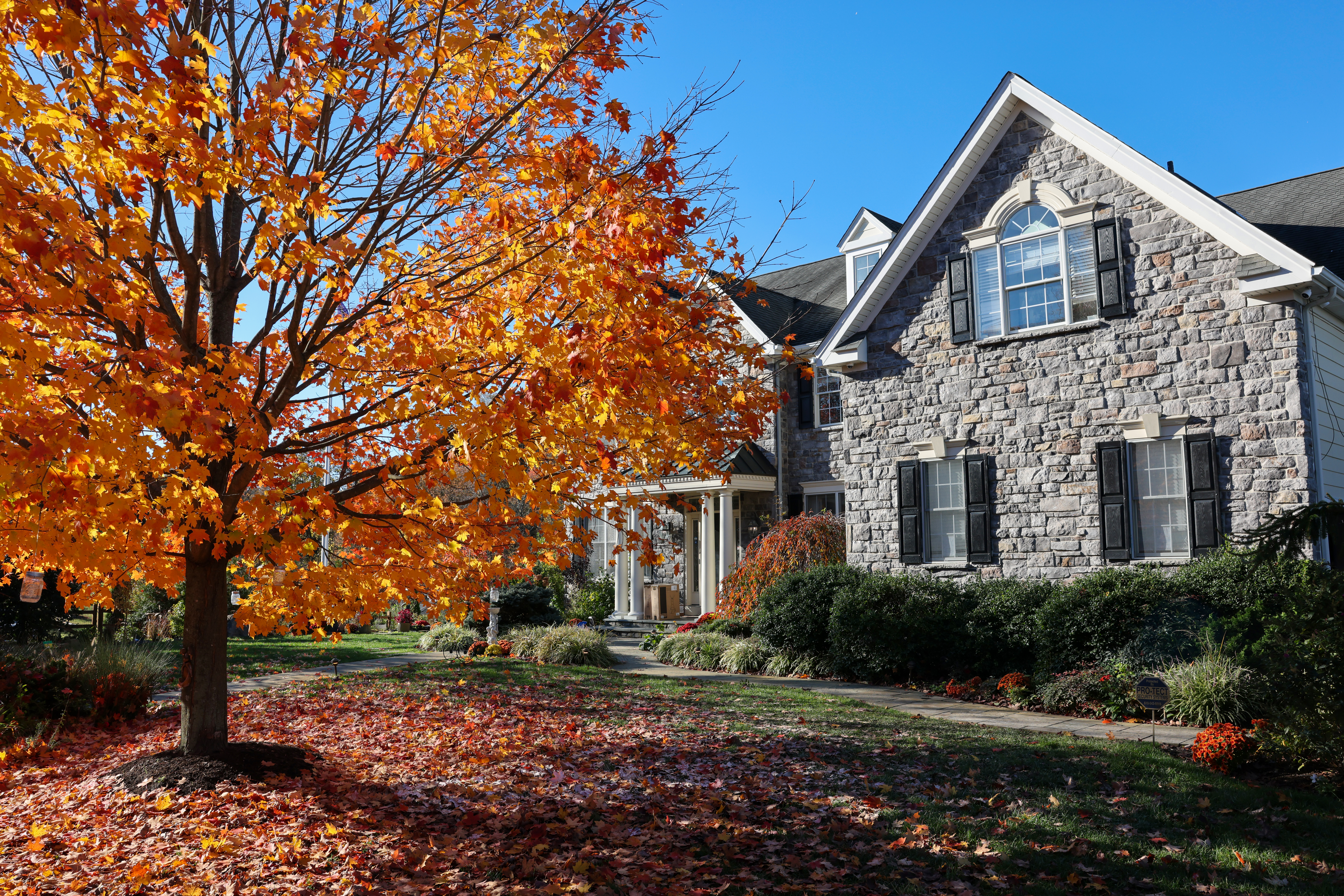 A stately stone home sits on a lawn covered in falling autumn leaves.