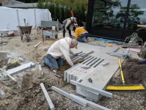 Two workers are installing rectangular stone pavers on a patio outside a building, with construction tools and materials scattered around them.