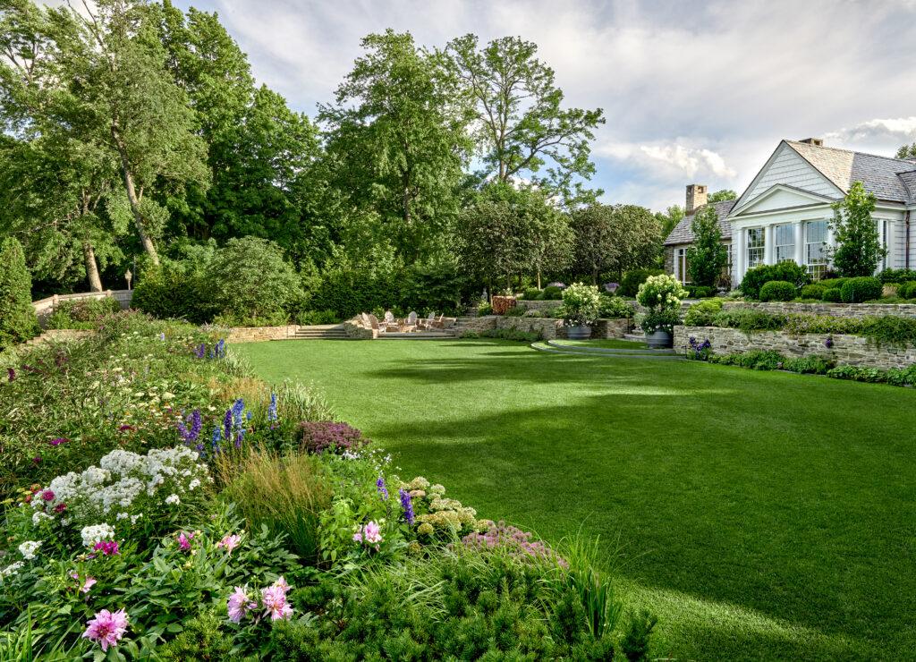A well-maintained lawn with flower beds, stone retaining walls, potted plants, and a white house surrounded by trees and outdoor seating—created through a collaborative design process.