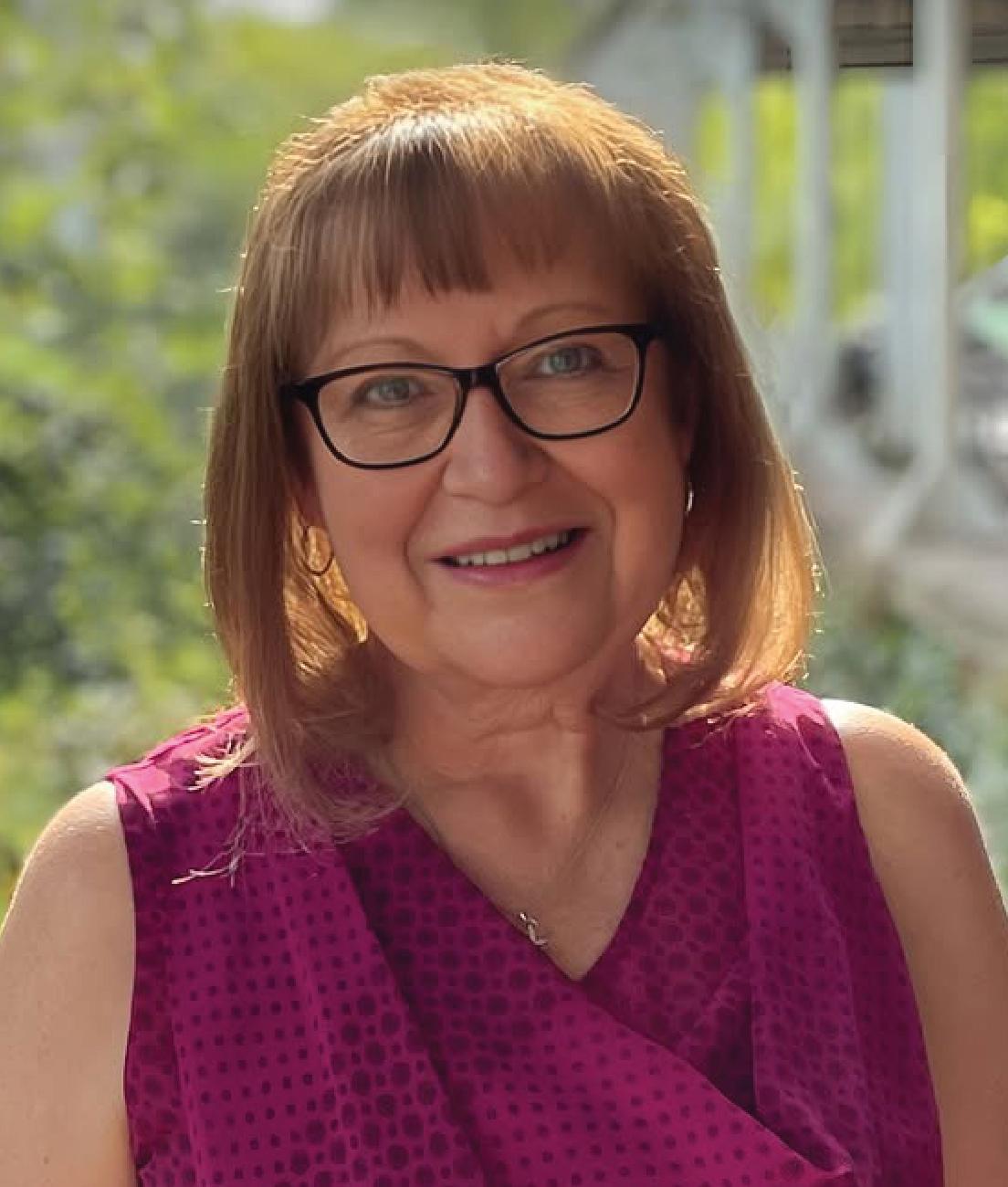 A woman with straight, shoulder-length light brown hair and glasses, wearing a sleeveless magenta top, smiles outdoors in natural light.