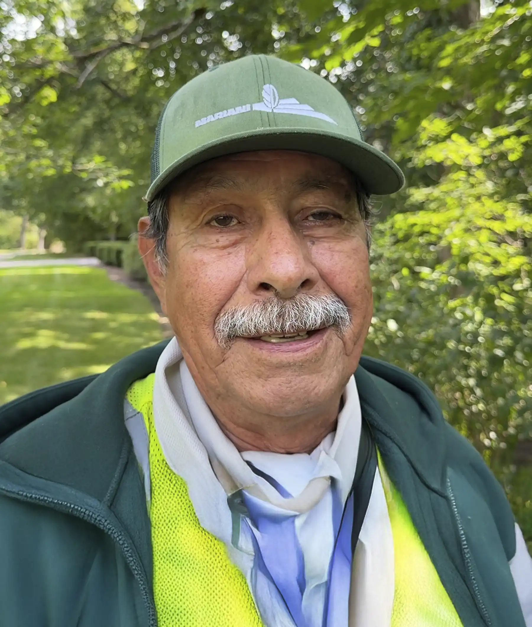 An older man with a mustache wearing a green cap, safety vest, and scarf stands outdoors in a park-like setting with trees in the background.