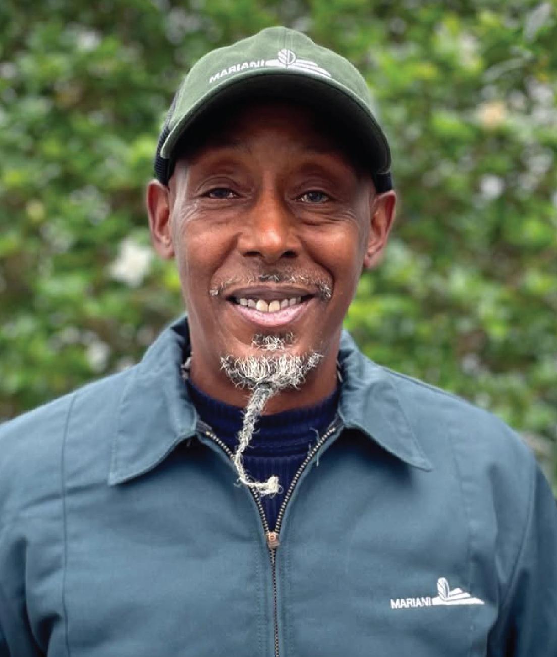 A man wearing a green cap and jacket stands outdoors, smiling. His beard is styled in a twisted, pointed shape. Green foliage is visible in the background.