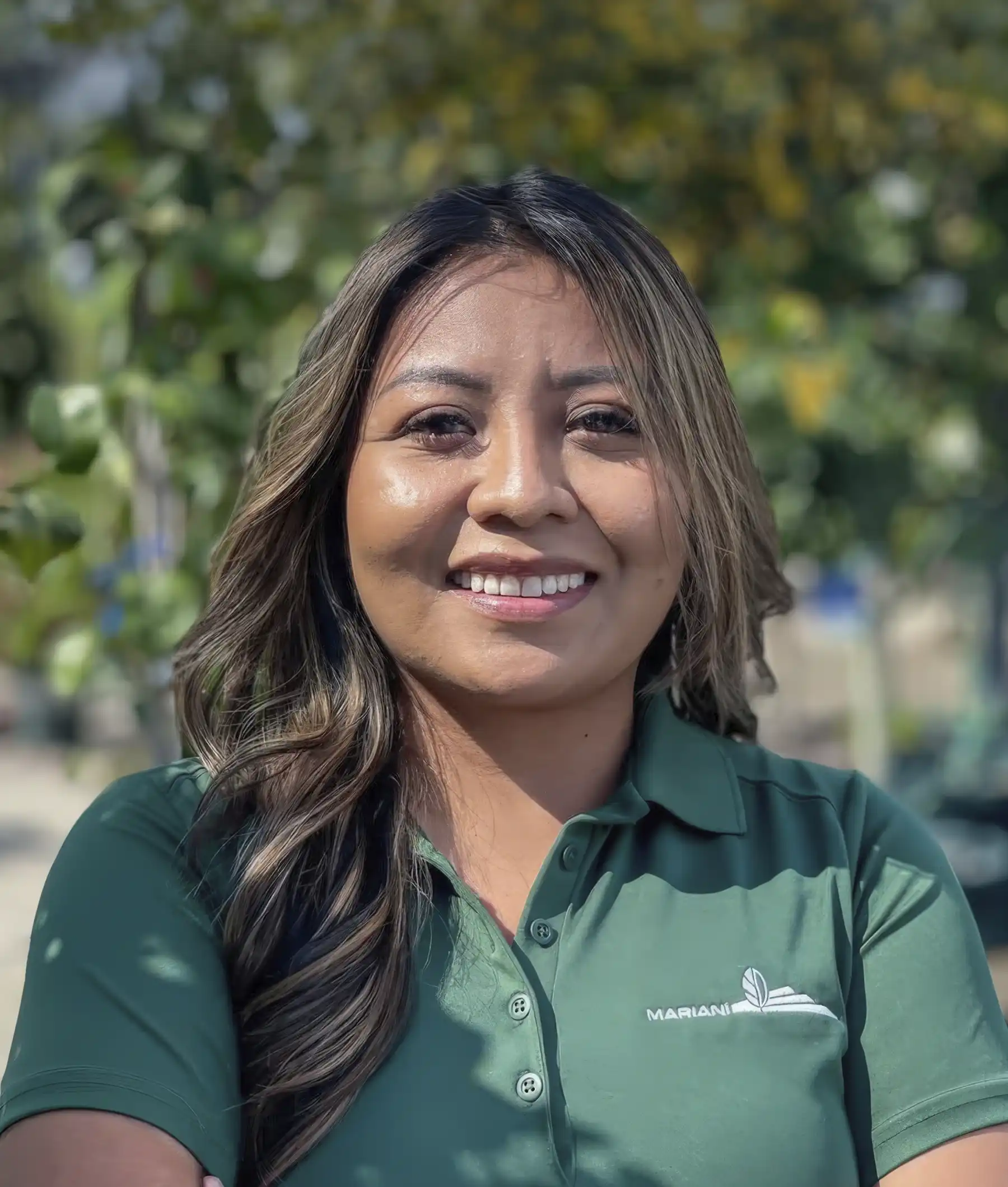 Woman with long brown hair wearing a green collared shirt stands outdoors, smiling at the camera with trees blurred in the background.