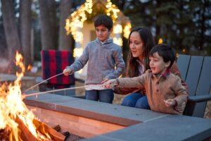 A woman and two young boys roast marshmallows over an outdoor fire pit, with string lights visible on a structure in the background.