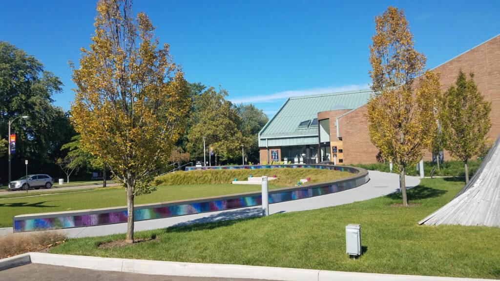 A curved pathway with colorful mosaic panels runs through a grassy area with trees, leading to a building with a green roof under a clear blue sky.
