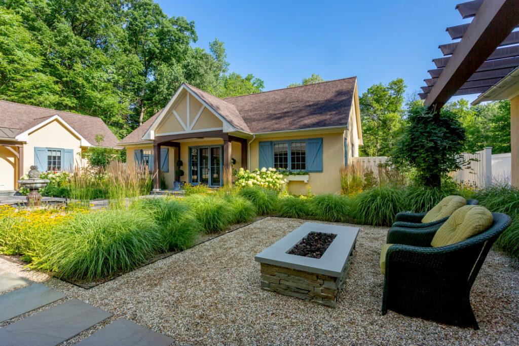 A cottage-style house with blue shutters and a landscaped garden, featuring outdoor wicker seating and a rectangular fire pit on gravel.