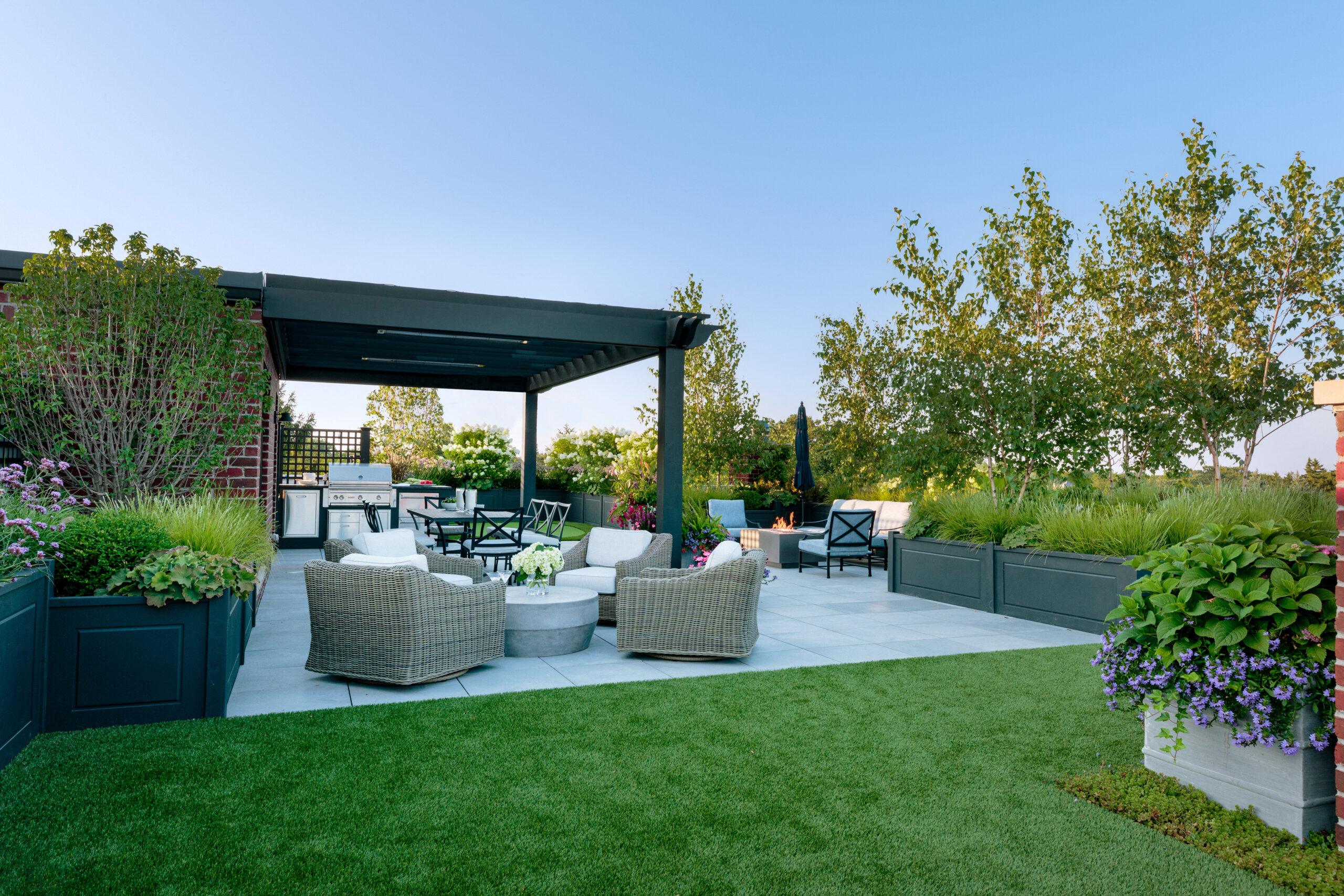 Outdoor patio with wicker furniture, a pergola, planters, and a lawn area; surrounded by greenery under a clear blue sky.