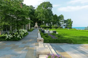 A landscaped backyard with stone patio, manicured lawn, garden beds, outdoor seating area, fireplace, and a view of trees and water in the background.