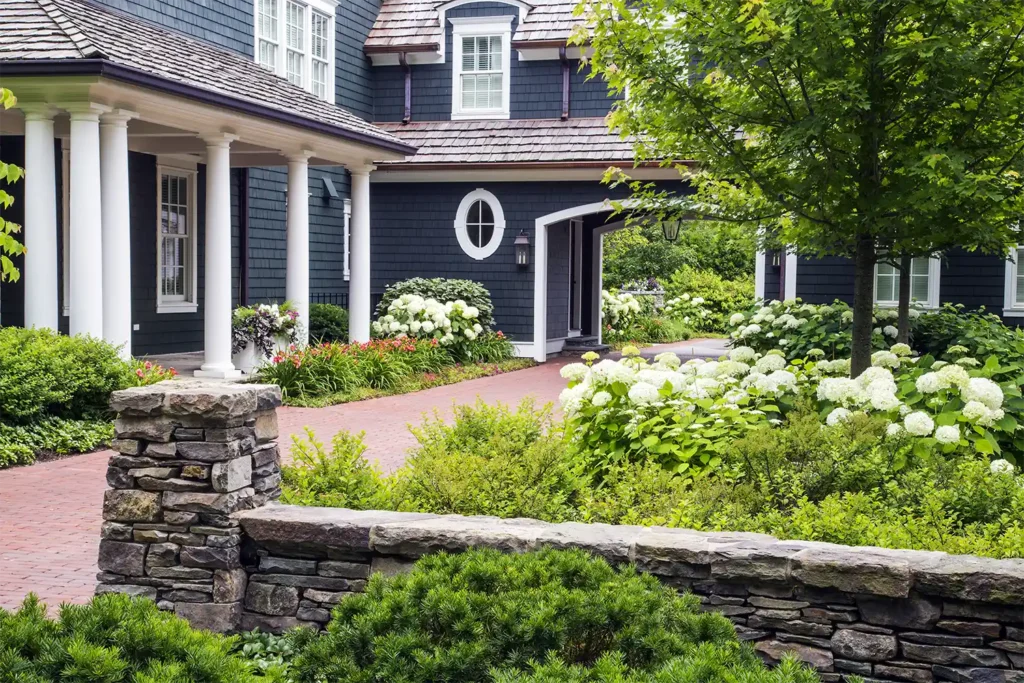 A landscaped front yard with a brick pathway, stone wall, green shrubs, blooming hydrangeas, and a dark blue house with white trim and columns in the background.