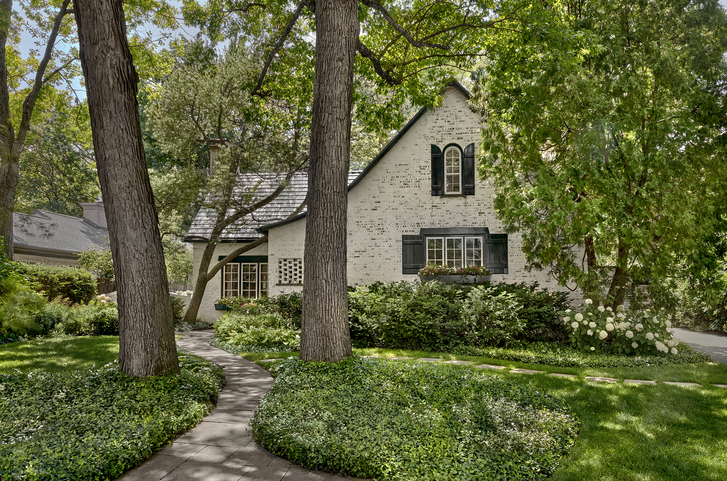 A white brick house with black shutters, arched windows, and a steep roof surrounded by mature trees, lush greenery, and a curved stone pathway.