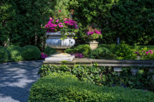 A stone planter filled with vibrant pink flowers sits beside a garden path, surrounded by manicured bushes and dense green foliage.