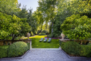 A landscaped garden with green lawn, three lounge chairs, stone planters, and surrounding trees and shrubs under clear daylight.