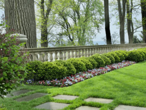 Neatly trimmed hedges and colorful flowers line a stone balustrade on a well-manicured lawn, with tall trees and a distant view in the background.