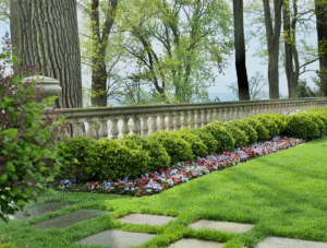 Neatly trimmed hedges and colorful flowers line a stone balustrade on a well-manicured lawn, with tall trees and a distant view in the background.