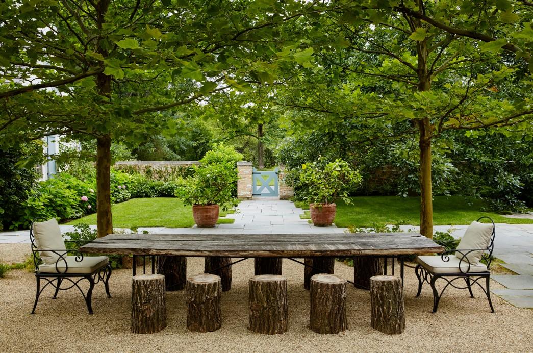 A rustic outdoor dining area features a long wooden table, log stools, cushioned chairs, and is shaded by two leafy trees in a lush garden setting.