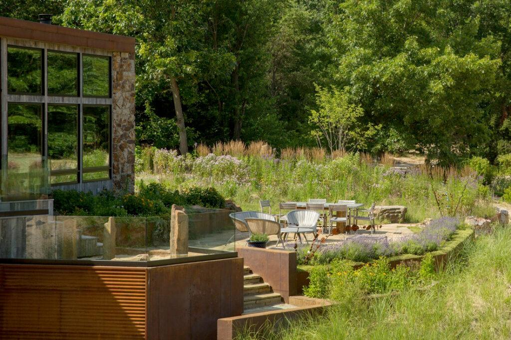 A modern house with large windows overlooks a landscaped garden featuring outdoor seating, a dining table, and glass railings surrounded by greenery.