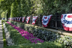 Decorative red, white, and blue bunting hung along a black iron fence above neatly trimmed hedges and bright pink flower beds.