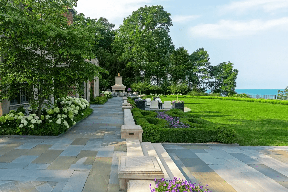 Expansive stone terrace with outdoor seating and fireplace facing a wide green lawn and lake bordered by mature trees.