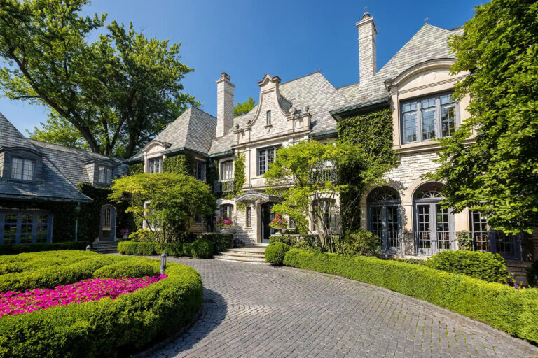 Grand stone mansion with ivy-covered façade, arched windows, manicured hedges, and a curved cobblestone driveway bordered by bright pink flowers.