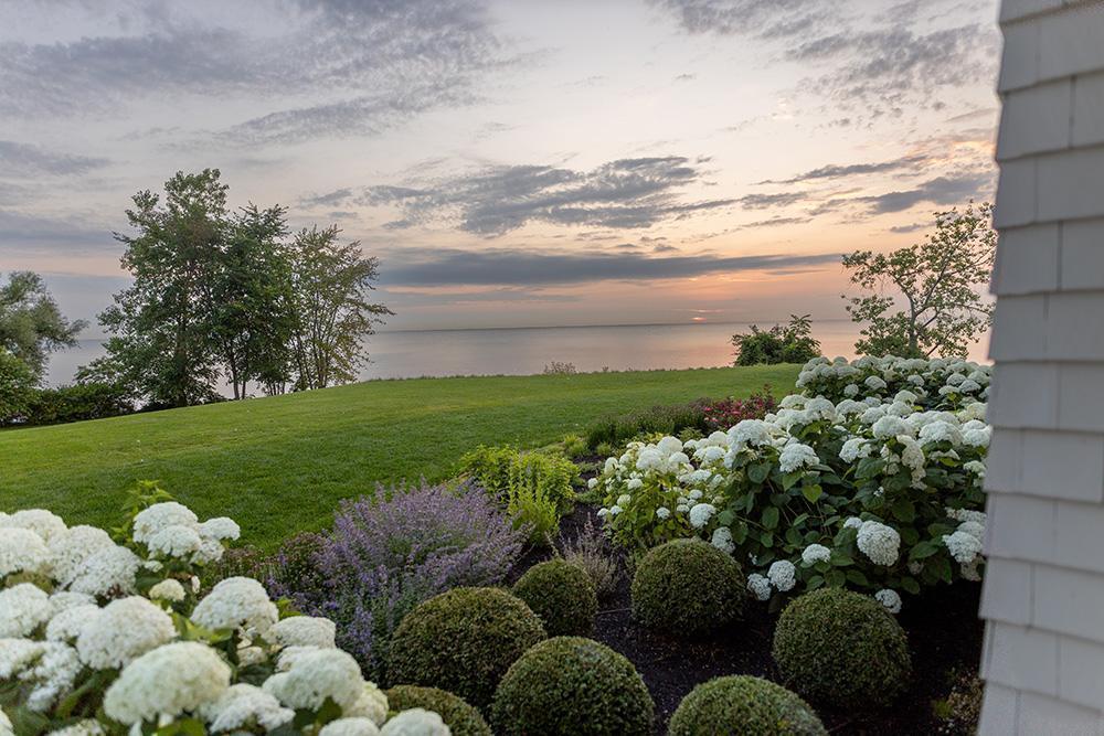 Lush backyard garden with white hydrangeas and trimmed shrubs leading to an open lawn overlooking a peaceful lake at dusk.