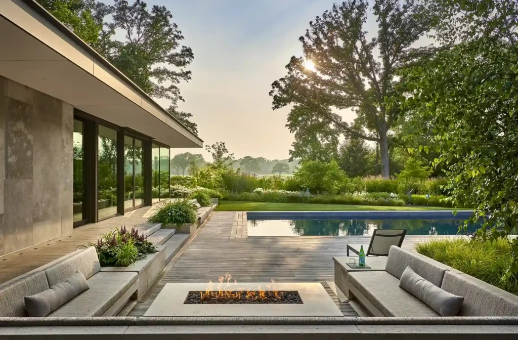 Modern patio with seating area and fire pit overlooking a rectangular pool and lush garden with large trees at sunrise.